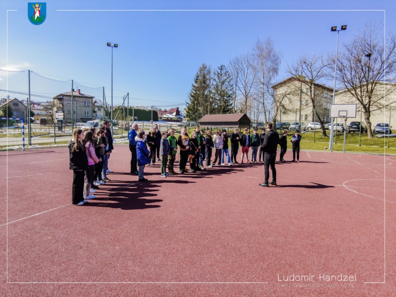 Radny Maciej Rogóż przemawia do dzieci na boisku sportowym.
